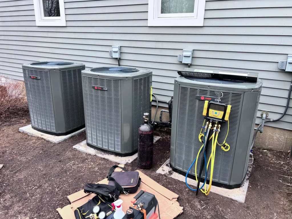 Three outdoor HVAC condenser units are installed beside a house, with the right unit undergoing maintenance as indicated by attached diagnostic gauges and tools laid out on the ground.