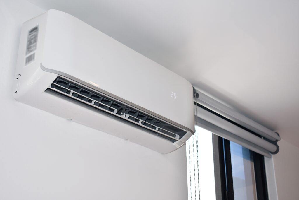 A wall-mounted ductless air conditioning unit is installed near a window with rolled-up blinds in a modern room.