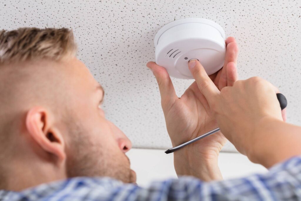 A man tests a carbon monoxide or smoke detector on a ceiling while holding a screwdriver.